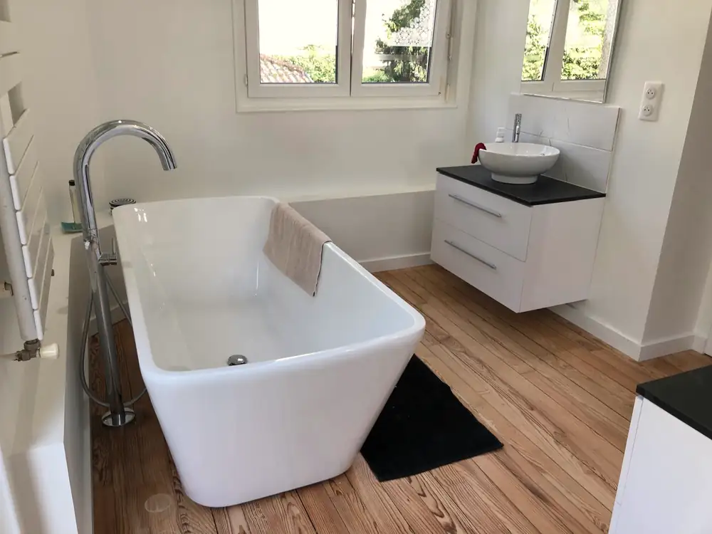 Baignoire îlot blanche posée sur parquet, robinetterie sur colonne et meuble vasque suspendu.