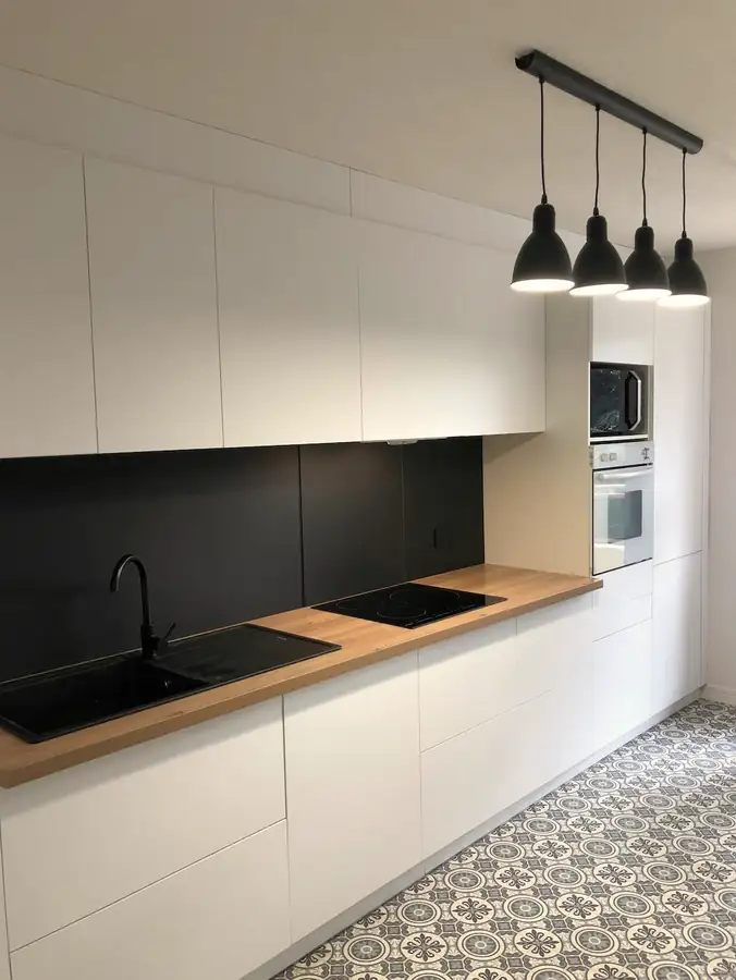 Cuisine blanche sans poignées, crédence noire, plan de travail bois et suspensions noires.