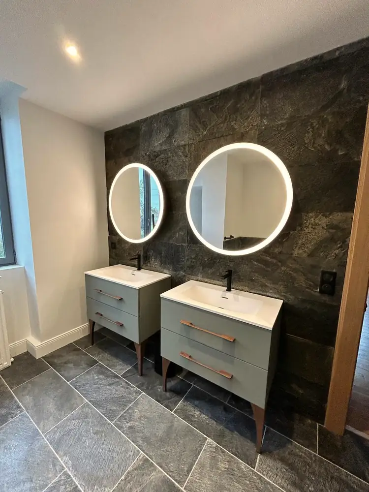Salle de bains avec double vasque, miroirs ronds rétroéclairés et carrelage effet pierre sombre.