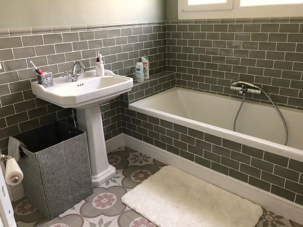 Salle de bains avec baignoire, faïence métro verte, lavabo sur colonne et sol en carreaux de ciment.