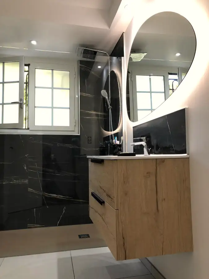 Salle de bains avec meuble vasque en chêne suspendu, miroirs ronds rétroéclairés et carrelage effet marbre noir.