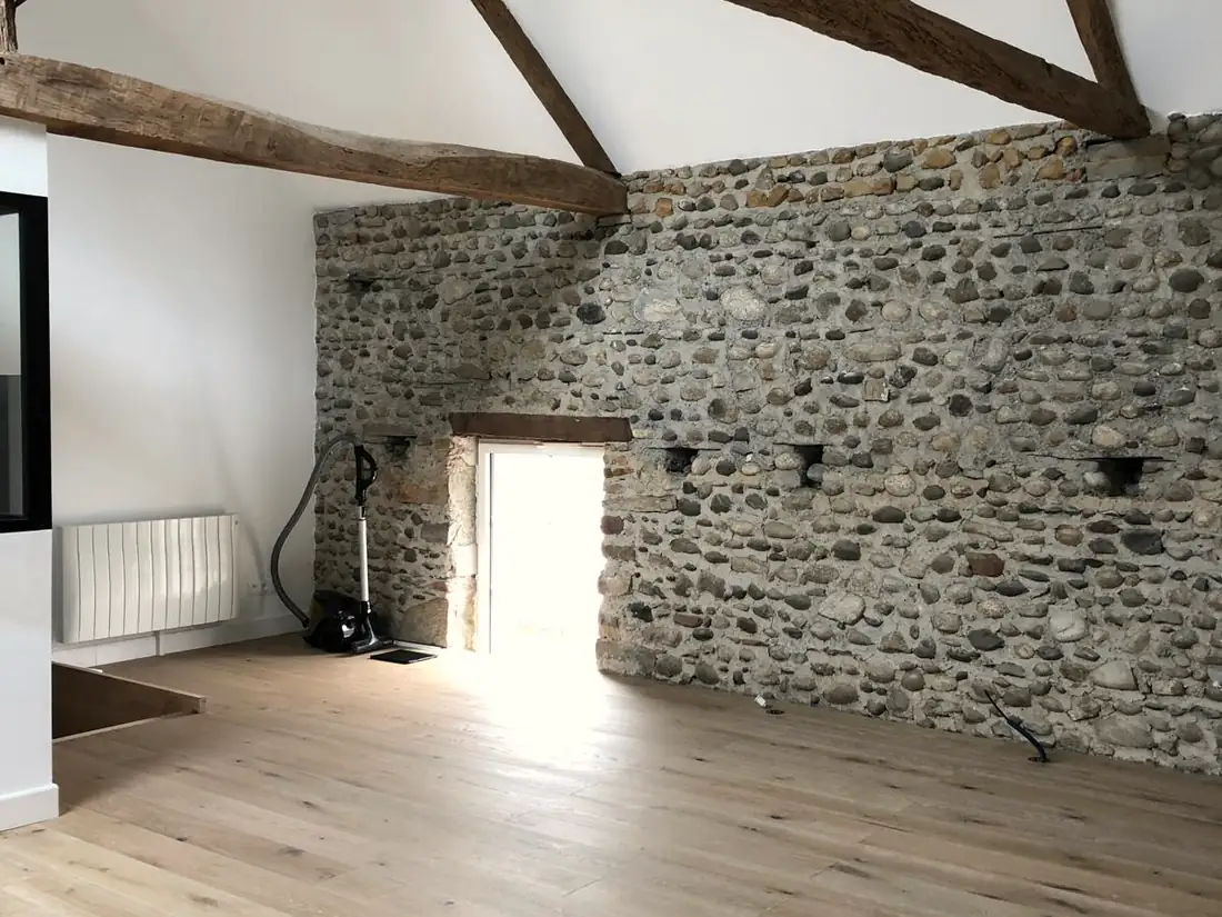 Grande pièce sous charpente apparente avec mur en galets et parquet clair.