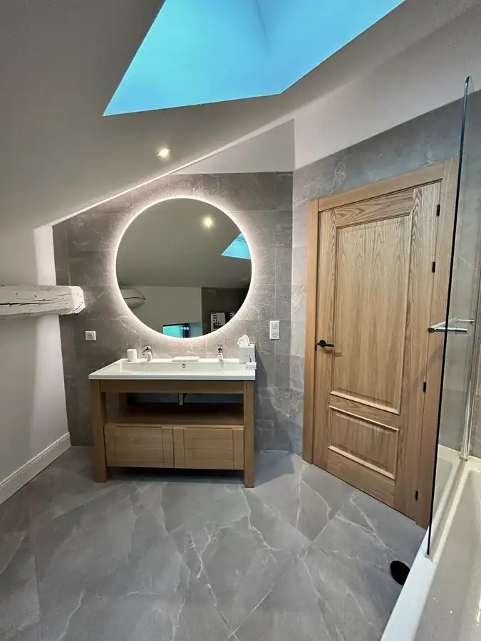 Salle de bains sous combles avec fenêtre de toit, miroir rond rétroéclairé et meuble vasque bois.
