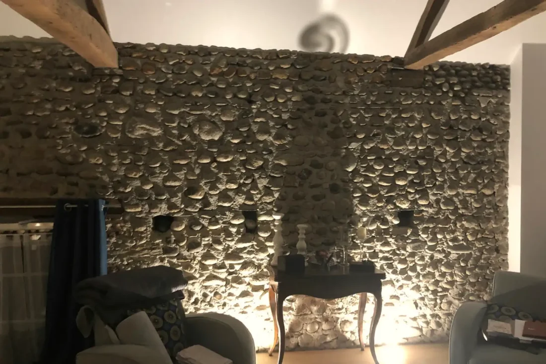 Séjour rénové sous charpente, mur en galets conservé et éclairé par des appliques.